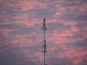 A telecommunications tower stands prominently against a backdrop of a vibrant, cloudy sky. The clouds are painted in hues of pink and purple, suggesting a sunset or sunrise.