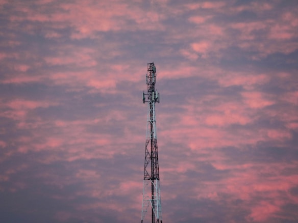 A telecommunications tower stands prominently against a backdrop of a vibrant, cloudy sky. The clouds are painted in hues of pink and purple, suggesting a sunset or sunrise.