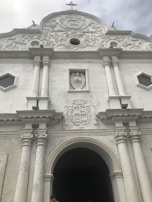 Facade of a white, ornate historical building with a large arched entrance flanked by two pairs of columns. Above the entrance is a sculpted crest and a rectangular relief featuring an angel. The building displays intricate architectural details and a small cross atop the roof.