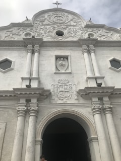 Facade of a white, ornate historical building with a large arched entrance flanked by two pairs of columns. Above the entrance is a sculpted crest and a rectangular relief featuring an angel. The building displays intricate architectural details and a small cross atop the roof.