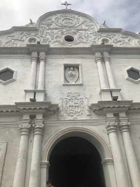 Facade of a white, ornate historical building with a large arched entrance flanked by two pairs of columns. Above the entrance is a sculpted crest and a rectangular relief featuring an angel. The building displays intricate architectural details and a small cross atop the roof.