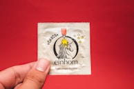 A condom packet is held by a hand against a plain red background. The packet features an illustration of a unicorn, with a yellow dripping substance above its head and several stars around it. Beneath the unicorn, the brand name 'einhorn' is printed along with the phrase 'make magic happen'.