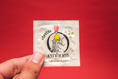 A condom packet is held by a hand against a plain red background. The packet features an illustration of a unicorn, with a yellow dripping substance above its head and several stars around it. Beneath the unicorn, the brand name 'einhorn' is printed along with the phrase 'make magic happen'.