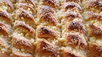 A close-up view of a freshly baked pastry with a golden-brown crust. The surface is textured with small granules, likely sugar or crumbs, and displays a pattern of overlapping layers. The pastry appears flaky and buttery.