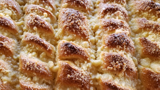 A close-up view of a freshly baked pastry with a golden-brown crust. The surface is textured with small granules, likely sugar or crumbs, and displays a pattern of overlapping layers. The pastry appears flaky and buttery.