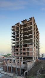 A tall, unfinished building stands amid a construction site. The structure consists of concrete frameworks and red brickwork. Scaffolding is visible on the sides; the surroundings include soil and construction materials.