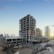 A multi-story building under construction is prominently positioned in the middle of a construction site, surrounded by dirt and construction materials. In the background, a few completed high-rise buildings are visible, suggesting an urban development area. The sky is clear with a hint of sunlight peeking through, indicating either dawn or dusk.