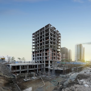 A multi-story building under construction is prominently positioned in the middle of a construction site, surrounded by dirt and construction materials. In the background, a few completed high-rise buildings are visible, suggesting an urban development area. The sky is clear with a hint of sunlight peeking through, indicating either dawn or dusk.