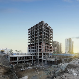 A multi-story building under construction is prominently positioned in the middle of a construction site, surrounded by dirt and construction materials. In the background, a few completed high-rise buildings are visible, suggesting an urban development area. The sky is clear with a hint of sunlight peeking through, indicating either dawn or dusk.