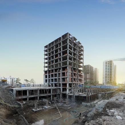 A multi-story building under construction is prominently positioned in the middle of a construction site, surrounded by dirt and construction materials. In the background, a few completed high-rise buildings are visible, suggesting an urban development area. The sky is clear with a hint of sunlight peeking through, indicating either dawn or dusk.