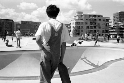 A black and white scene of an urban skate park with several people engaging in various activities. A person in the foreground, wearing overalls and holding a skateboard, looks towards the skate bowls. In the background, skaters are actively riding and performing tricks, while bystanders watch. The backdrop features modern apartment buildings under a partly cloudy sky.
