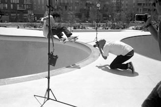 A black and white photograph captures the moment a skateboarder performs an aerial trick above a skate bowl, with a photographer kneeling nearby to capture the action. Several lighting stands are set up around the bowl, indicating a professional photo shoot. In the background, there are people sitting and standing, watching the action, against the backdrop of urban buildings.