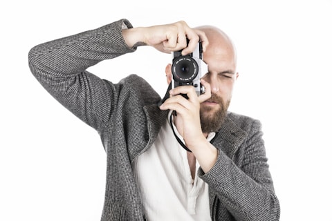 A person with a shaved head and beard is holding up a camera to their face, as if taking a photo. They are dressed in a patterned blazer over a light-colored shirt. The background is white, creating a high-contrast, minimalist look.