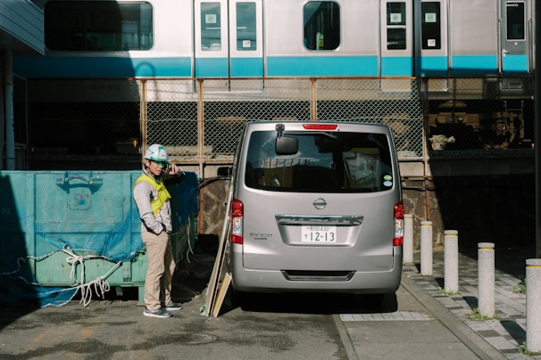 A construction worker wearing a hard hat and safety vest stands next to a silver van parked near construction materials. In the background, a train on elevated tracks passes by a chain-link fence.