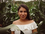 A young woman with long dark hair and a white off-shoulder top is standing amidst tall green corn plants. The greenery surrounds her, with lush leaves framing the foreground. Her expression is serene and calm.