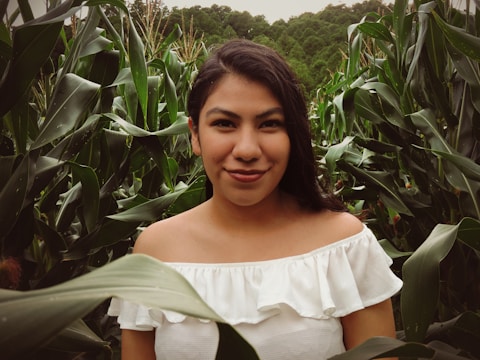 A young woman with long dark hair and a white off-shoulder top is standing amidst tall green corn plants. The greenery surrounds her, with lush leaves framing the foreground. Her expression is serene and calm.