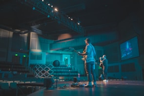 Two musicians perform on stage in a dimly lit auditorium. One is playing a guitar while the other is singing into a microphone. The setting includes a large display screen, empty seats, and stage lighting rigs overhead, contributing to a concert-like atmosphere.