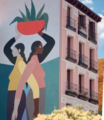 A mural painted on the side of a building featuring two stylized figures in profile standing back-to-back. One figure is holding an orange pot with green plants on her head. The building has a pink façade with several balconies and black railings. The scene is set against a bright blue sky.
