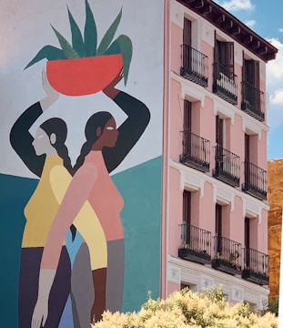 A mural painted on the side of a building featuring two stylized figures in profile standing back-to-back. One figure is holding an orange pot with green plants on her head. The building has a pink façade with several balconies and black railings. The scene is set against a bright blue sky.