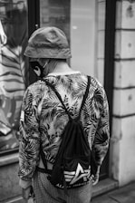 A person wearing a bucket hat, a face mask, and a patterned jacket with leaf motifs. They carry a drawstring bag with a visible logo of an 'A' and the words 'Alpha Industries.' The individual's back is facing the camera, and they are standing near a storefront with a reflection of foliage patterns.