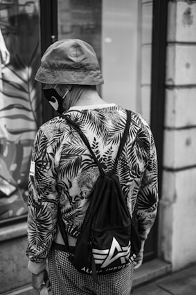 A person wearing a bucket hat, a face mask, and a patterned jacket with leaf motifs. They carry a drawstring bag with a visible logo of an 'A' and the words 'Alpha Industries.' The individual's back is facing the camera, and they are standing near a storefront with a reflection of foliage patterns.