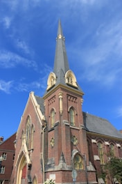 A tall, historic brick church with a prominent spire reaching towards a clear blue sky. The building features Gothic architectural elements with pointed arches, lancet windows, and decorative stonework.