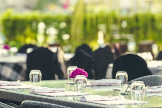 A set of outdoor tables and chairs are arranged with elegant glassware and napkins. In the foreground, a small bouquet of purple flowers adds a pop of color. The background is lush with greenery, and the lighting suggests a bright, sunny day.