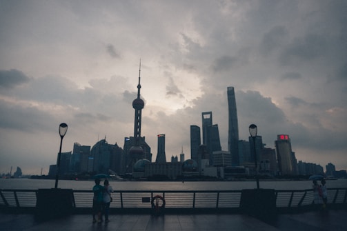 Cloudy skyline of a modern city with a prominent TV tower centered against dramatic clouds. Two silhouetted figures holding umbrellas stand on a waterfront promenade, with street lamps in view.