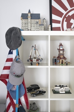 A shelf displaying various model structures and objects, including a detailed castle model on top, a space vehicle with figures, a clock tower, several scale model cars, and a model of the White House. Hanging on the side are several caps and an American flag.