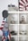 A shelf displaying various model structures and objects, including a detailed castle model on top, a space vehicle with figures, a clock tower, several scale model cars, and a model of the White House. Hanging on the side are several caps and an American flag.