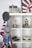A shelf displaying various model structures and objects, including a detailed castle model on top, a space vehicle with figures, a clock tower, several scale model cars, and a model of the White House. Hanging on the side are several caps and an American flag.