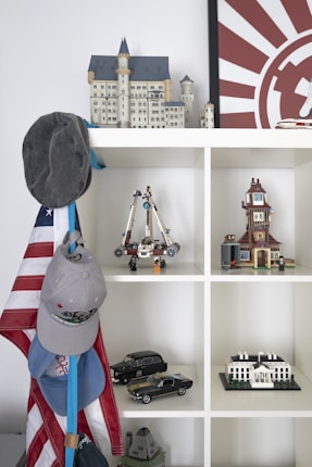 A shelf displaying various model structures and objects, including a detailed castle model on top, a space vehicle with figures, a clock tower, several scale model cars, and a model of the White House. Hanging on the side are several caps and an American flag.