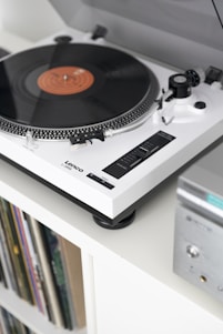 A vintage-style turntable with a spinning vinyl record is mounted on a white shelf. The turntable features a sleek, modern design with a white and black color scheme. Below the turntable, there is a collection of vinyl records stored in a shelf. An audio receiver with a silver finish is partially visible on the right side.