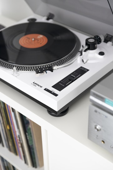 A vintage-style turntable with a spinning vinyl record is mounted on a white shelf. The turntable features a sleek, modern design with a white and black color scheme. Below the turntable, there is a collection of vinyl records stored in a shelf. An audio receiver with a silver finish is partially visible on the right side.