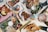 An assortment of dishes including crispy fried fish with lemon slices, breaded chicken, various salads, and garnished appetizers. The table is styled with checkered paper and wooden utensils, alongside cans of soda and a glass with a clear beverage.