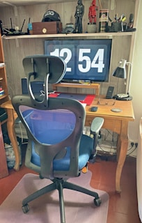 A home office setup with a large digital clock display on a monitor showing the time 12:54. In front of the monitor is a wooden desk with various items including a lamp, a laptop, eyeglasses, and a smartphone. A modern office chair with a blue mesh back is positioned in front of the desk. Shelving above the desk holds figurines, a cap with a Star Wars logo, books, and decorative items.