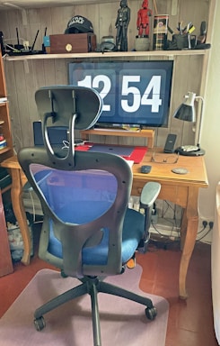 A home office setup with a large digital clock display on a monitor showing the time 12:54. In front of the monitor is a wooden desk with various items including a lamp, a laptop, eyeglasses, and a smartphone. A modern office chair with a blue mesh back is positioned in front of the desk. Shelving above the desk holds figurines, a cap with a Star Wars logo, books, and decorative items.