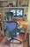 A home office setup with a large digital clock display on a monitor showing the time 12:54. In front of the monitor is a wooden desk with various items including a lamp, a laptop, eyeglasses, and a smartphone. A modern office chair with a blue mesh back is positioned in front of the desk. Shelving above the desk holds figurines, a cap with a Star Wars logo, books, and decorative items.
