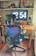 A home office setup with a large digital clock display on a monitor showing the time 12:54. In front of the monitor is a wooden desk with various items including a lamp, a laptop, eyeglasses, and a smartphone. A modern office chair with a blue mesh back is positioned in front of the desk. Shelving above the desk holds figurines, a cap with a Star Wars logo, books, and decorative items.