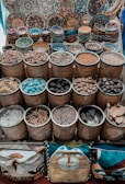 Handcrafted, colorful bowls and plates with intricate designs are displayed on a surface, along with bamboo baskets filled with a variety of spices, nuts, and other natural materials. Cushions with artistic designs are placed at the bottom of the display.