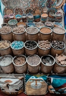 Handcrafted, colorful bowls and plates with intricate designs are displayed on a surface, along with bamboo baskets filled with a variety of spices, nuts, and other natural materials. Cushions with artistic designs are placed at the bottom of the display.