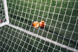 A close-up view of a football pitch, with a focus on two bright orange markers near the goal net. The green artificial turf is marked with a white line, and the net adds a texture to the scene.