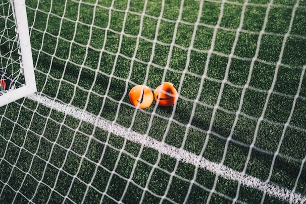 A close-up view of a football pitch, with a focus on two bright orange markers near the goal net. The green artificial turf is marked with a white line, and the net adds a texture to the scene.