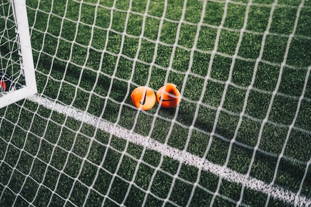 A close-up view of a football pitch, with a focus on two bright orange markers near the goal net. The green artificial turf is marked with a white line, and the net adds a texture to the scene.