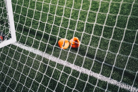 A close-up view of a football pitch, with a focus on two bright orange markers near the goal net. The green artificial turf is marked with a white line, and the net adds a texture to the scene.
