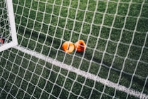 A close-up view of a football pitch, with a focus on two bright orange markers near the goal net. The green artificial turf is marked with a white line, and the net adds a texture to the scene.