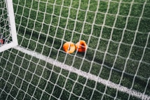 A close-up view of a football pitch, with a focus on two bright orange markers near the goal net. The green artificial turf is marked with a white line, and the net adds a texture to the scene.