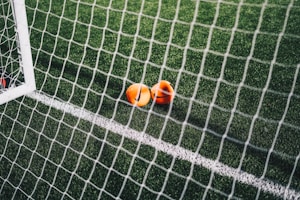 A close-up view of a football pitch, with a focus on two bright orange markers near the goal net. The green artificial turf is marked with a white line, and the net adds a texture to the scene.