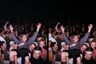 A large group of people gathered in a celebratory atmosphere, many smiling and raising their hands. The central figure is hoisted above the crowd, suggesting a lively, energetic event.