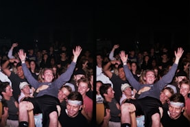 A large group of people gathered in a celebratory atmosphere, many smiling and raising their hands. The central figure is hoisted above the crowd, suggesting a lively, energetic event.